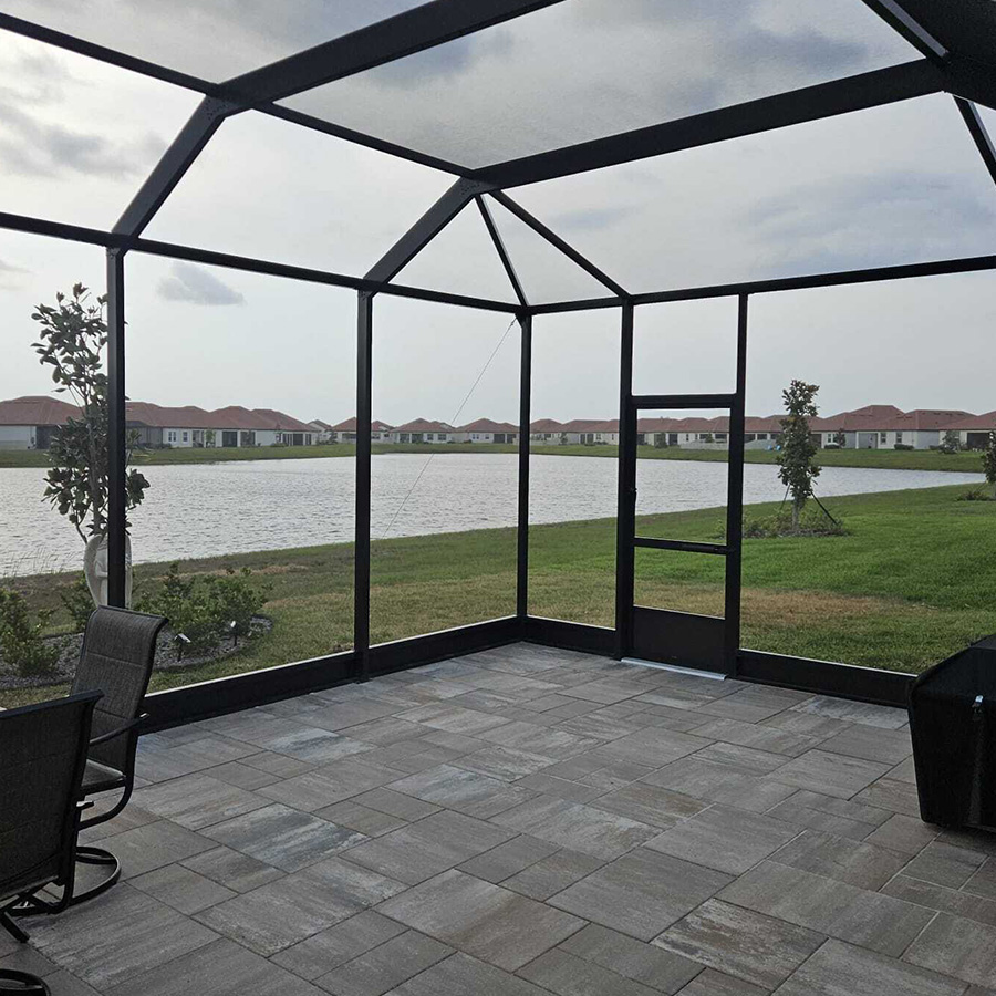 Interior view of a black-framed screen enclosure shows a paved patio that looks out over a calm body of water and a row of distant houses. The structure features a peaked roof design and a screen door that leads to the grassy backyard.