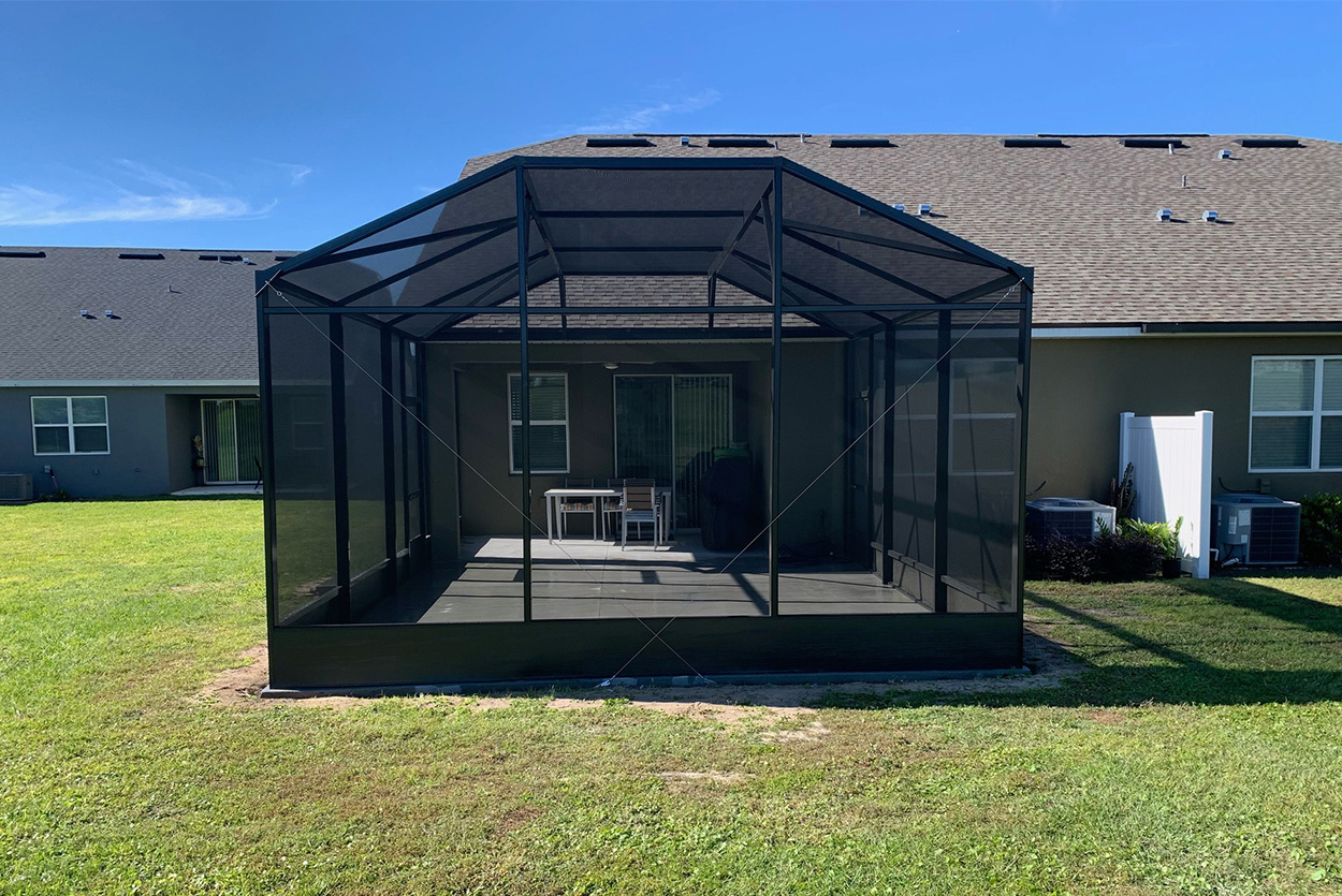 A black-framed screen enclosure is attached to the rear of a dark-colored, one-story home. The structure features a gabled screen roof and sits on a concrete patio, containing a dining table and chairs. The enclosure is situated on a flat green lawn, with a white fence and neighboring houses visible in the background.