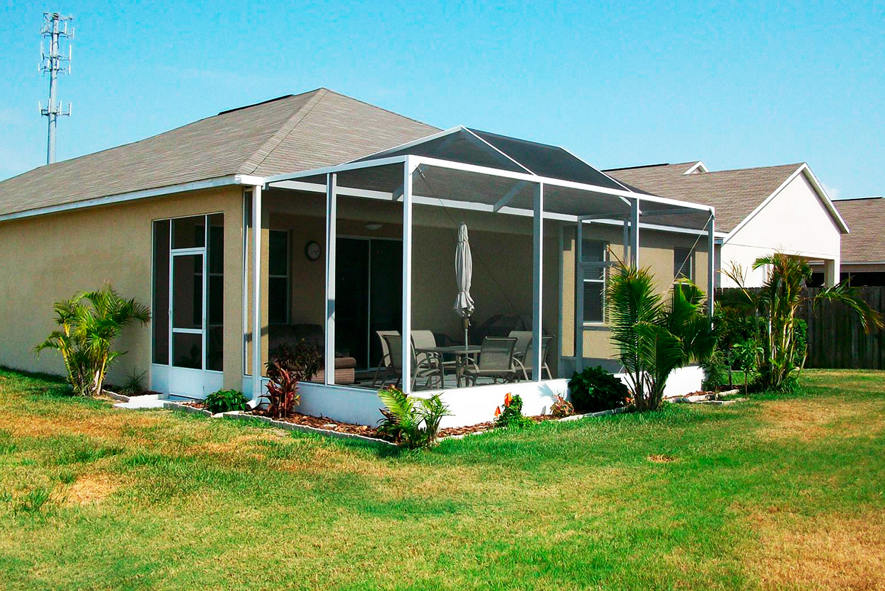 A white-framed screen enclosure is attached to the rear of a tan, one-story home. The enclosure features a peaked screen roof and a solid white base wall, housing a patio dining set and an umbrella.