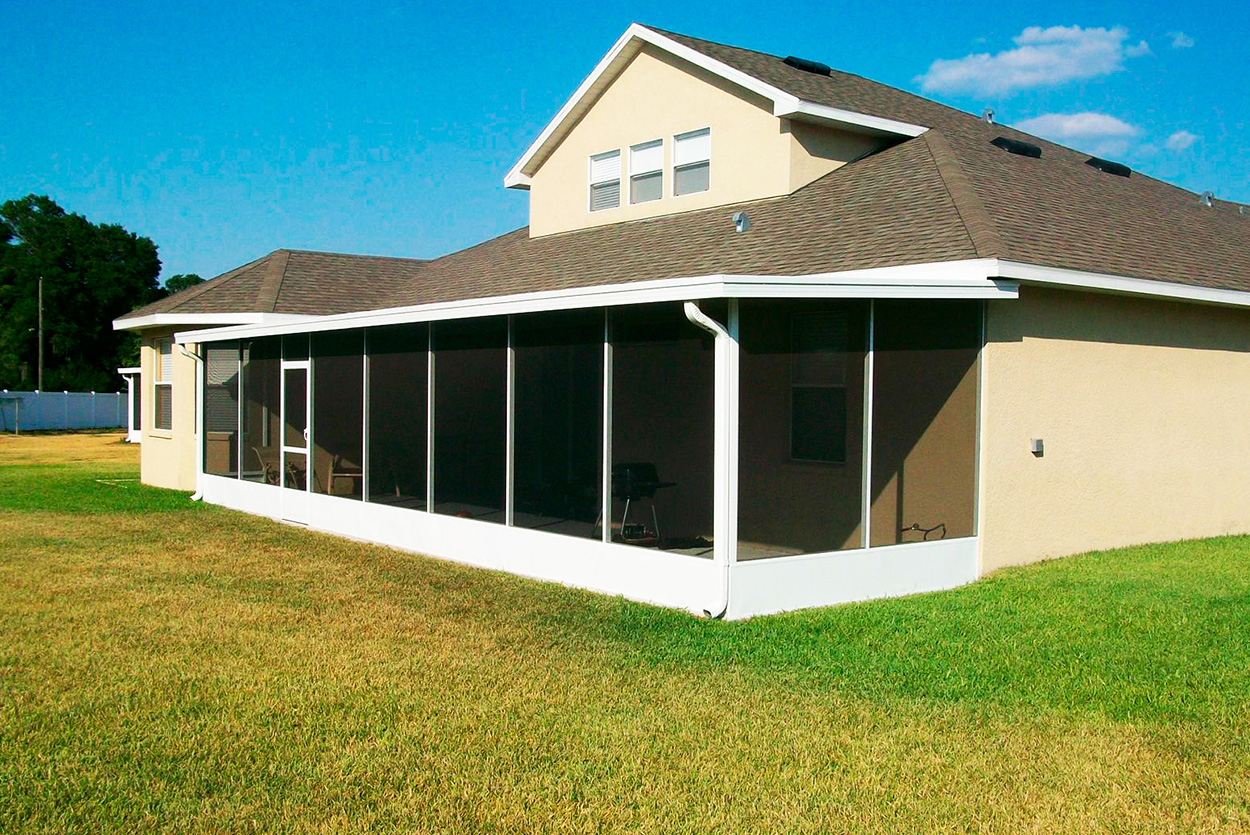 A white-framed screen enclosure is attached to the rear of a tan, two-story home. The enclosure features a solid white base wall and multiple vertical screen panels, extending along the side of the house over a grassy lawn.