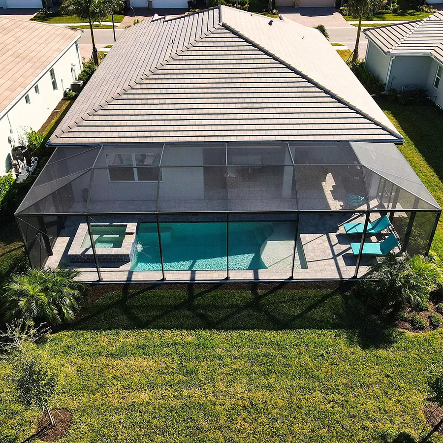 A rectangular swimming pool and integrated spa sit within a black mesh screen enclosure behind a tan house with a tiled roof. Teal lounge chairs rest on the stone paver deck beside the blue water.