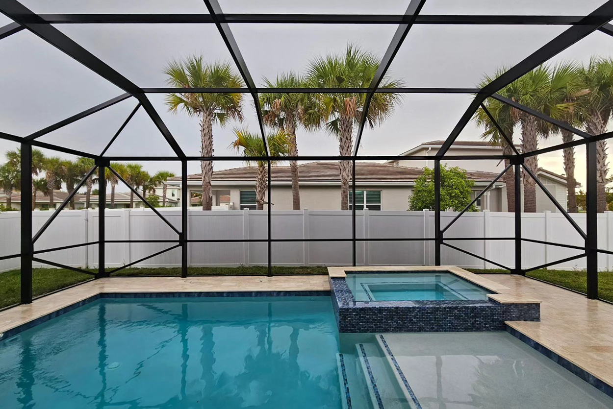 Backyard swimming pool features a clean, integrated design with a connected spa and light-toned stone decking. A black screen enclosure covers the entire area, providing protection while offering clear views of the palm trees.