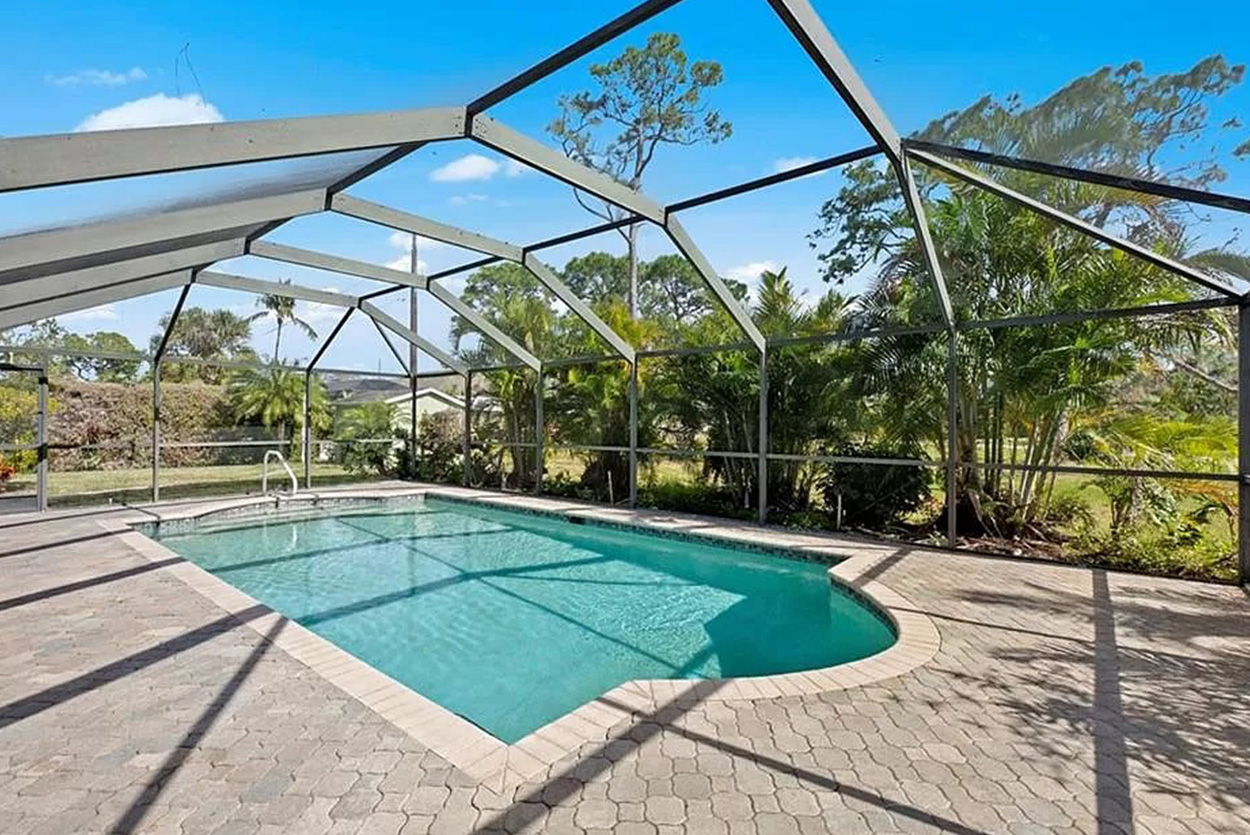 Pool is completely enclosed by a large mesh screen structure, providing a sheltered swimming environment while maintaining a clear view of the surrounding tropical landscape, palm trees, and neighboring homes under a bright sky.
